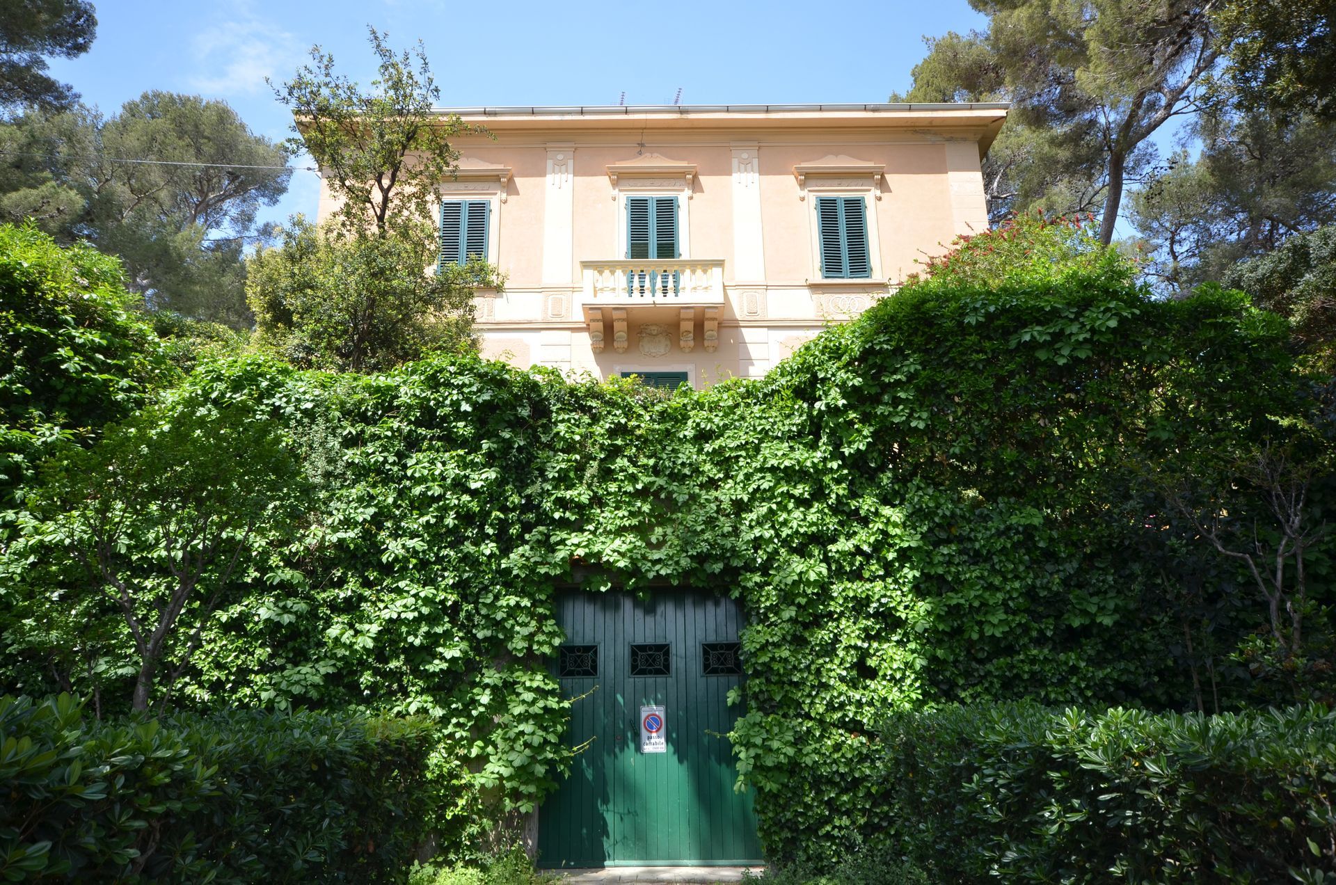 Casa color crema a due piani dietro una fitta edera verde con un cancello verde e alberi