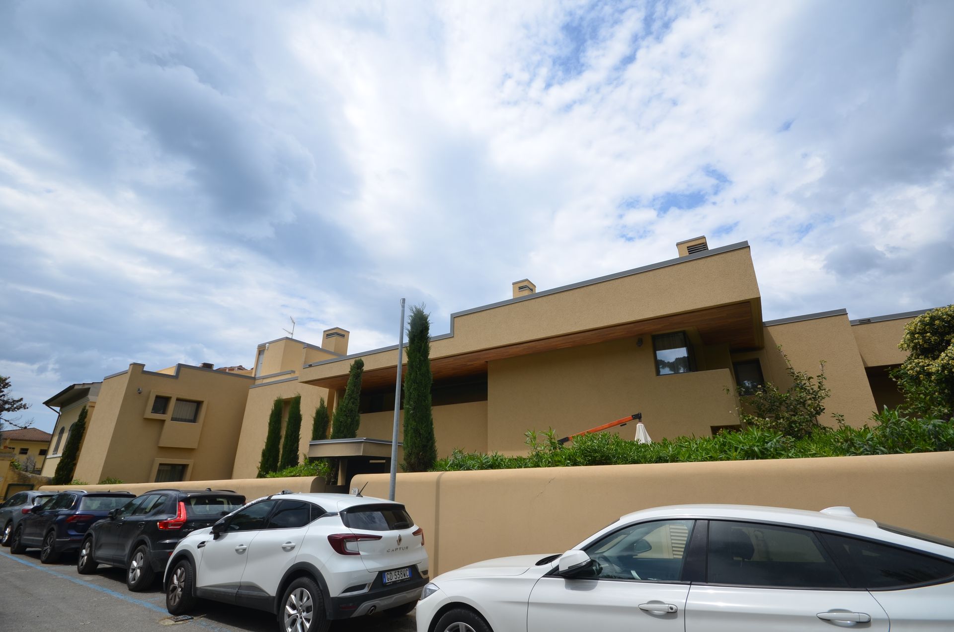 Un palazzo color sabbia con auto parcheggiate lungo una strada sotto un cielo azzurro nuvoloso.