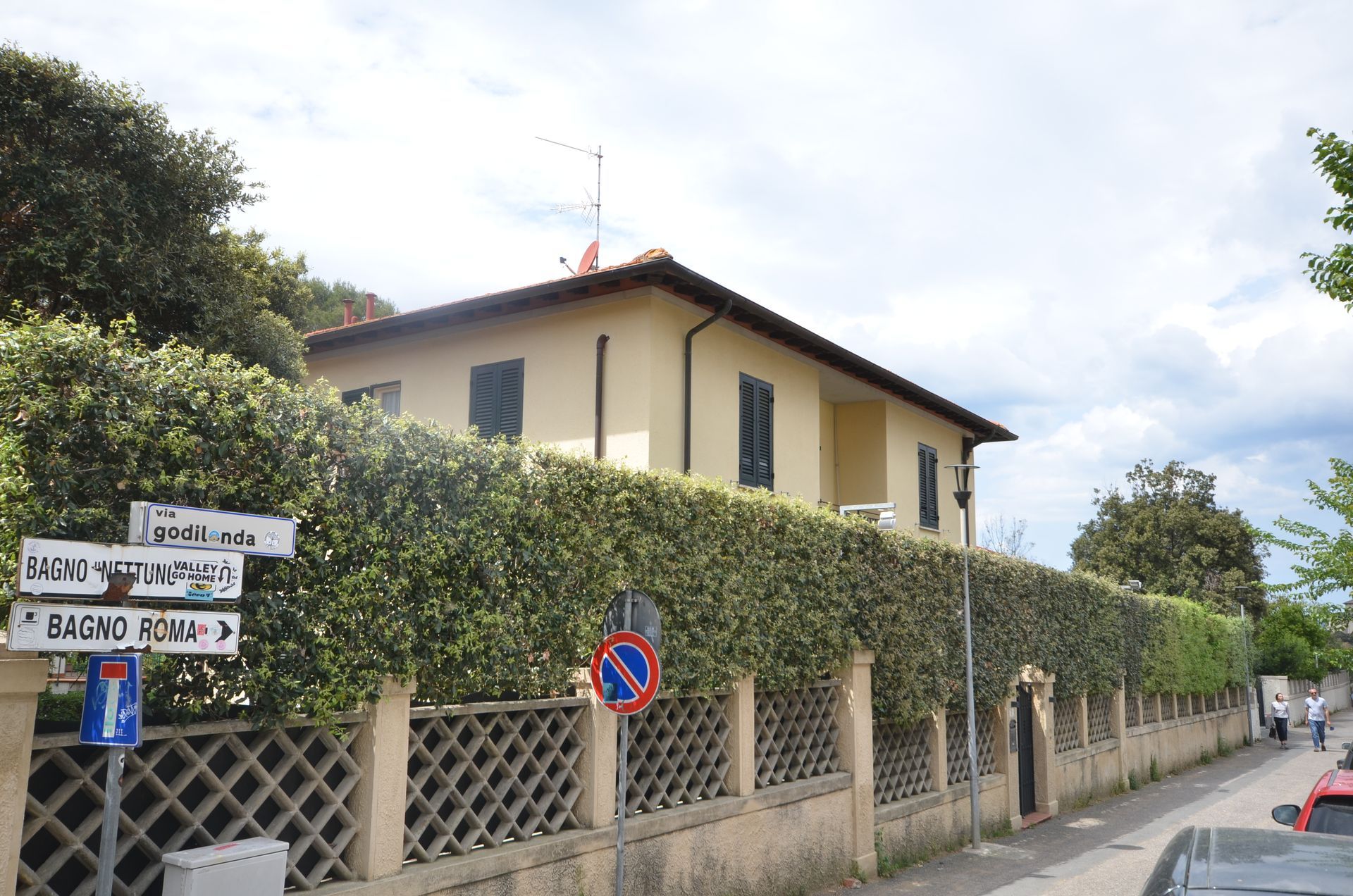 Angolo di strada con una casa color crema dietro un'alta siepe, segnali stradali e un marciapiede tranquillo in una giornata nuvolosa.