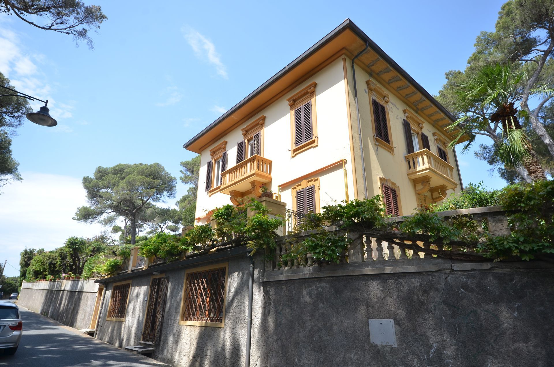 Una grande casa color crema e giallo dietro un muro di pietra, con alberi e cielo azzurro in una giornata di sole.