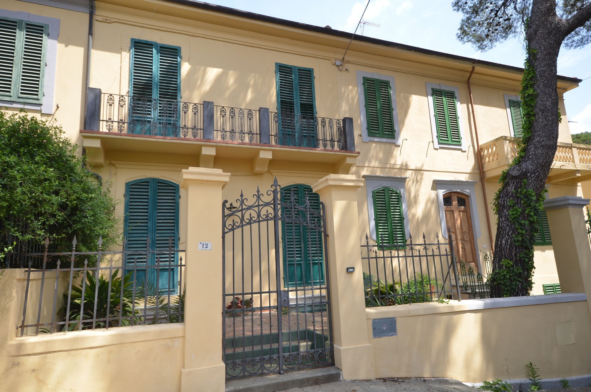 Casa beige a due piani con persiane blu e cancello in ferro battuto, vista dalla strada.
