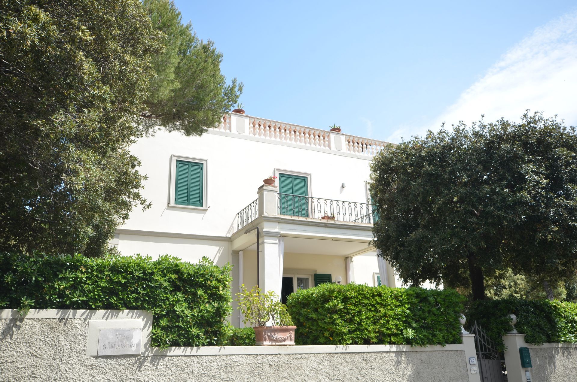 Casa bianca a due piani con persiane verdi e balcone, incorniciata da alberi e siepi sotto un cielo azzurro limpido.