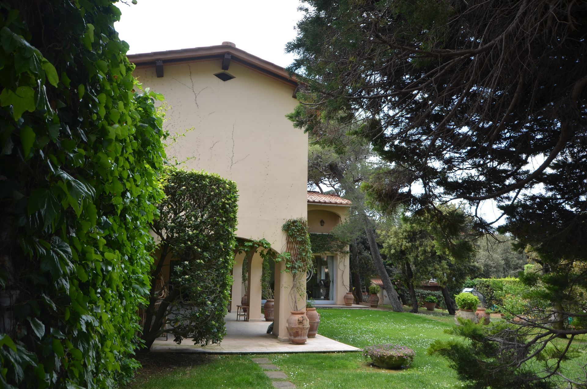Sentiero nel giardino accanto a una casa in stucco color crema con colonne ricoperte di edera e vegetazione lussureggiante.