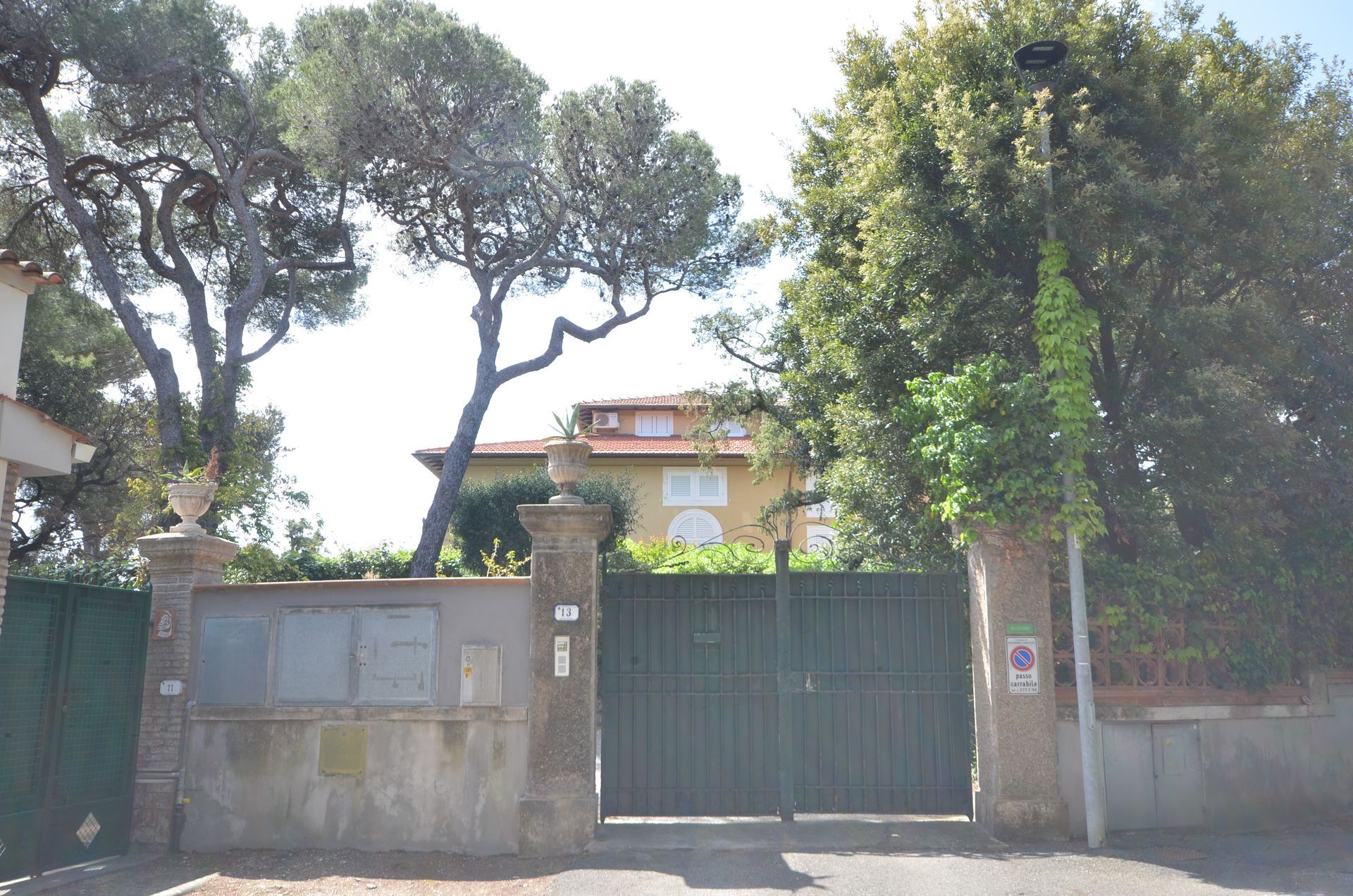 Ingresso della casa fiancheggiato da alberi, con cancelli verdi e muri in pietra, illuminato da una luce solare intensa.