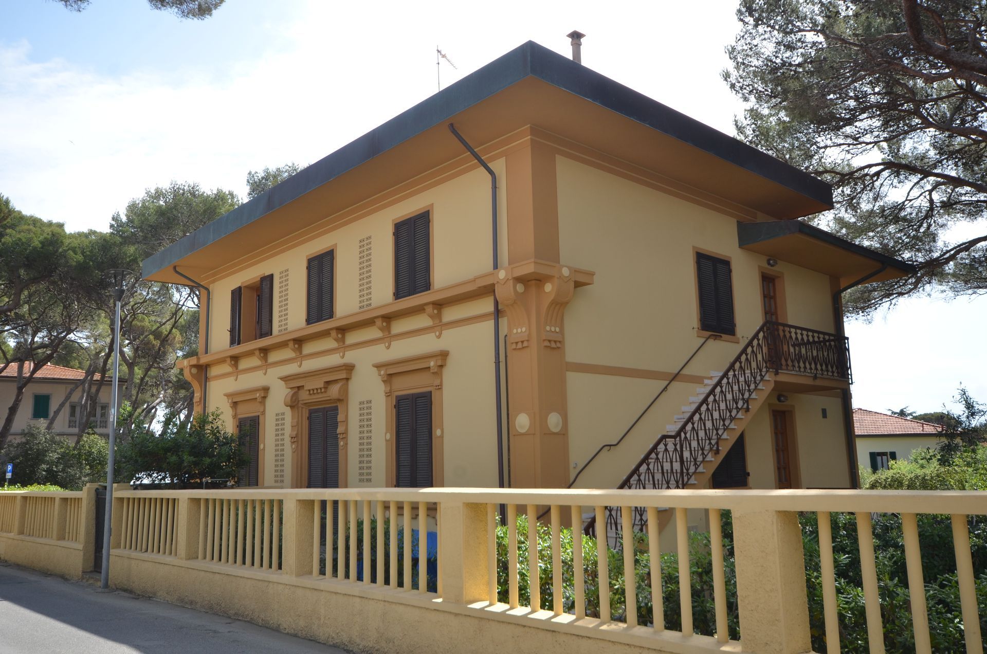 Edificio beige a due piani con tetto scuro, scale esterne e recinzione bianca sul davanti.