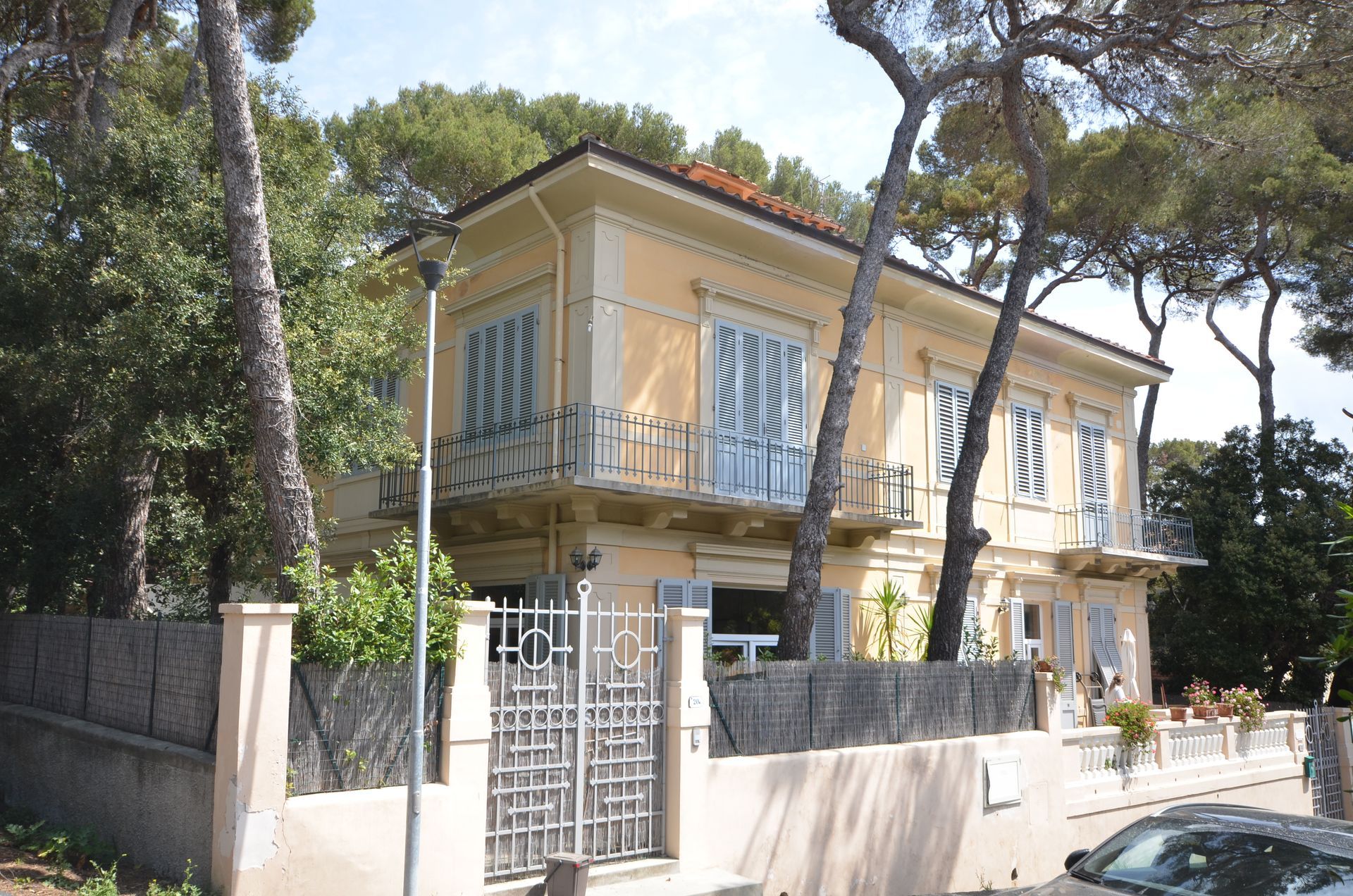 Villa gialla a due piani dietro un muro di pietra, con persiane blu e alti pini di fronte.