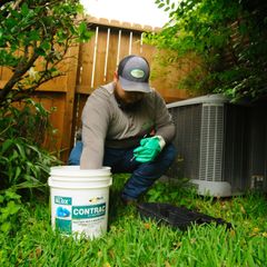 Man in hat and gloves leans into a bucket next to an AC unit in a grassy yard.