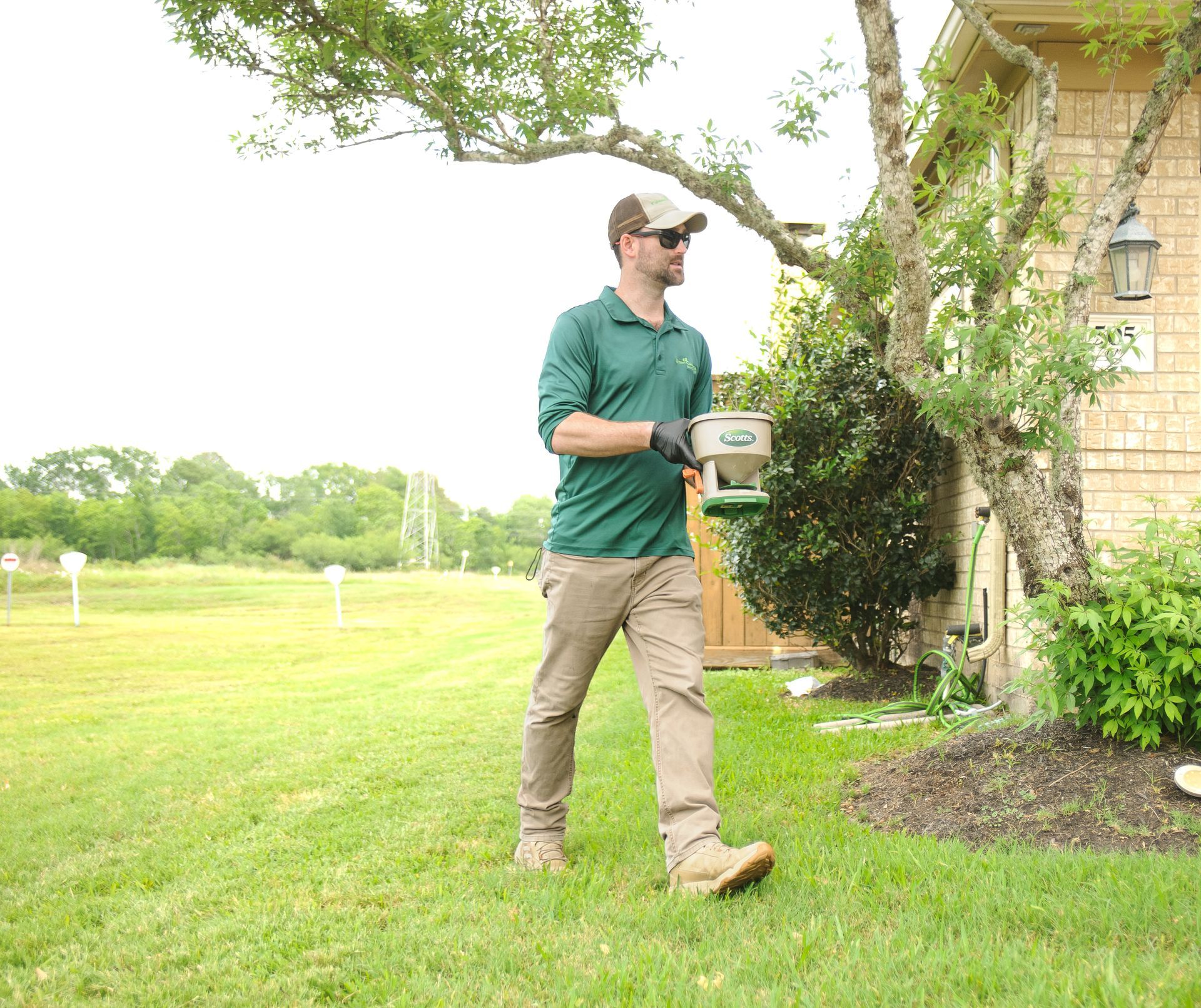 Man in green shirt and hat, carrying a spreader across a grassy yard.