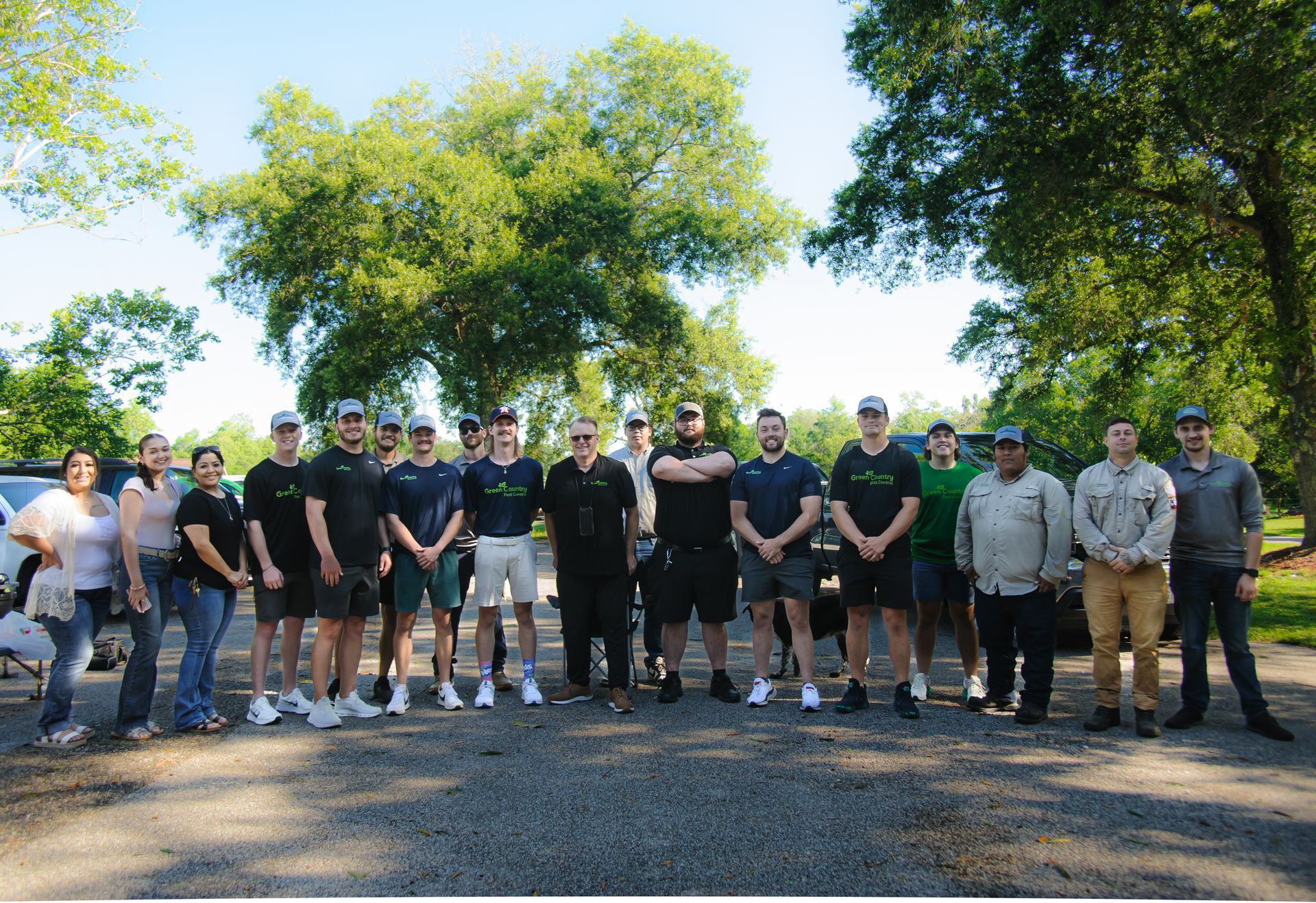Group of people in front of trees posing for a photo, wearing company shirts, outside in sunlight.