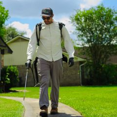 Pest control technician walking on a sidewalk, wearing a backpack sprayer, gray pants, and sunglasses.
