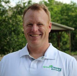 Man wearing a white polo shirt smiles, Green Country Pest Control logo visible. Outdoors, trees in background.