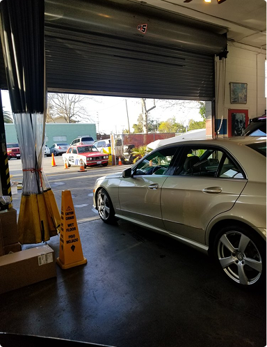 Silver sedan inside a garage with a partially open roll-up door, looking out to a street scene.  | Hudson Automotive
