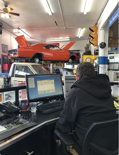 Person at a computer inside an auto repair shop with a red car lifted in the background  | Hudson Automotive