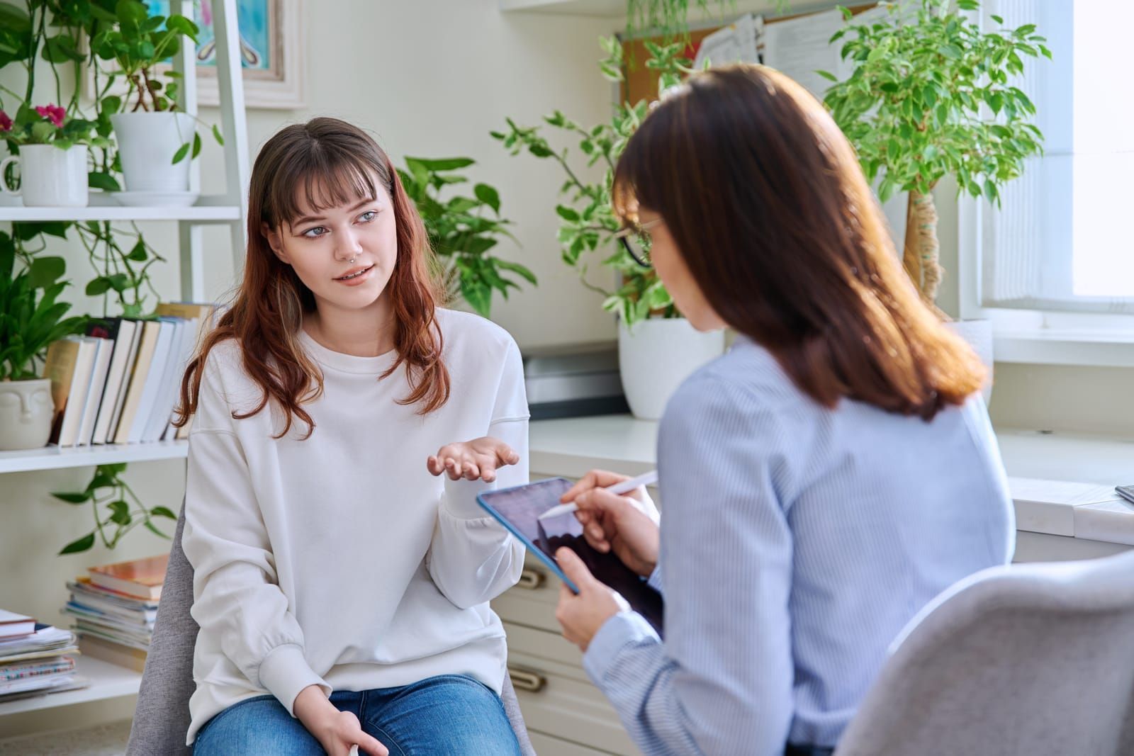 Two women talking in a bright living room, one holding a tablet and the other gesturing on a couch