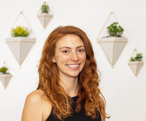 A woman with red hair is smiling in front of a wall with hanging potted plants.