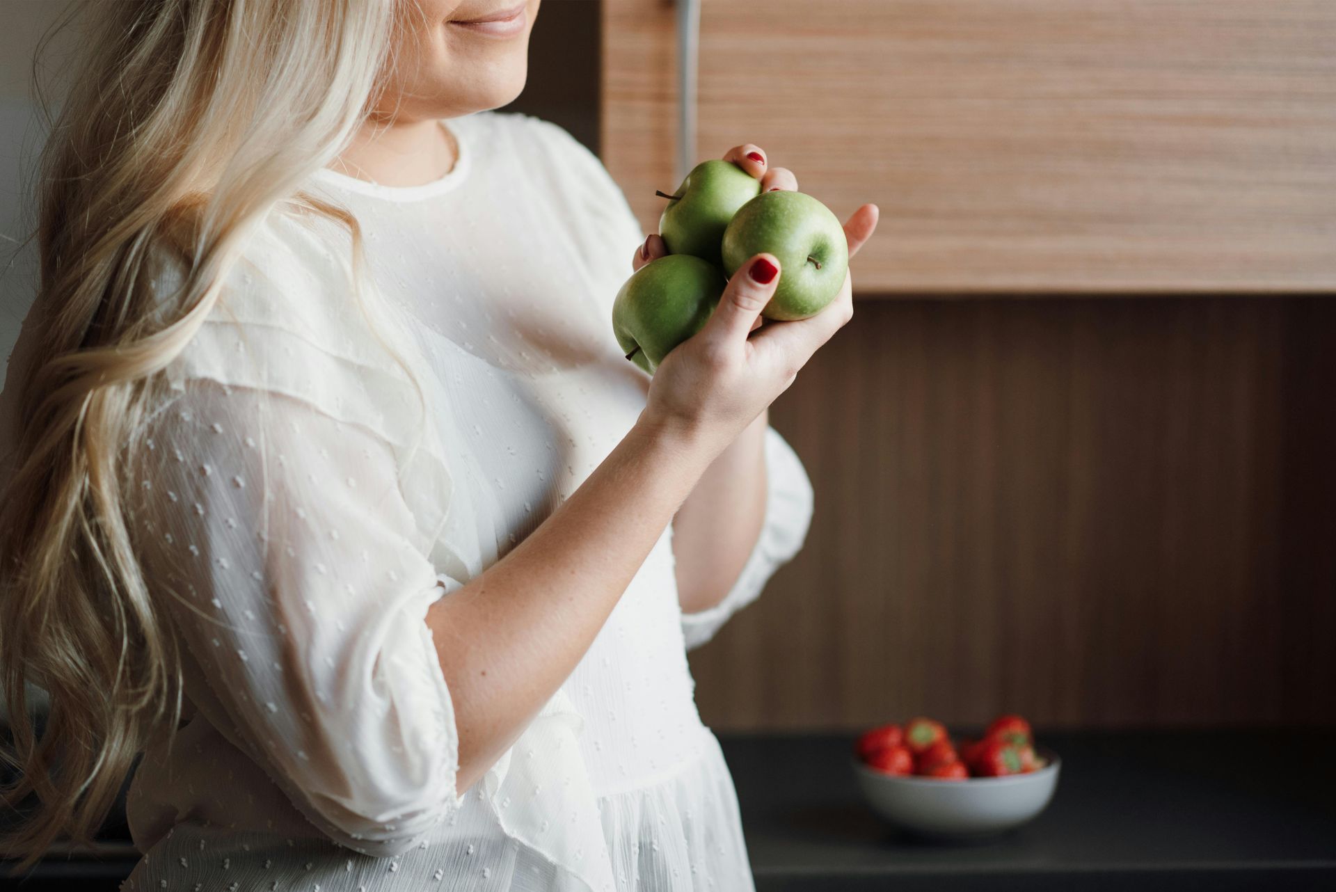 Person holding green apples in a kitchen with a bowl of strawberries on the counter