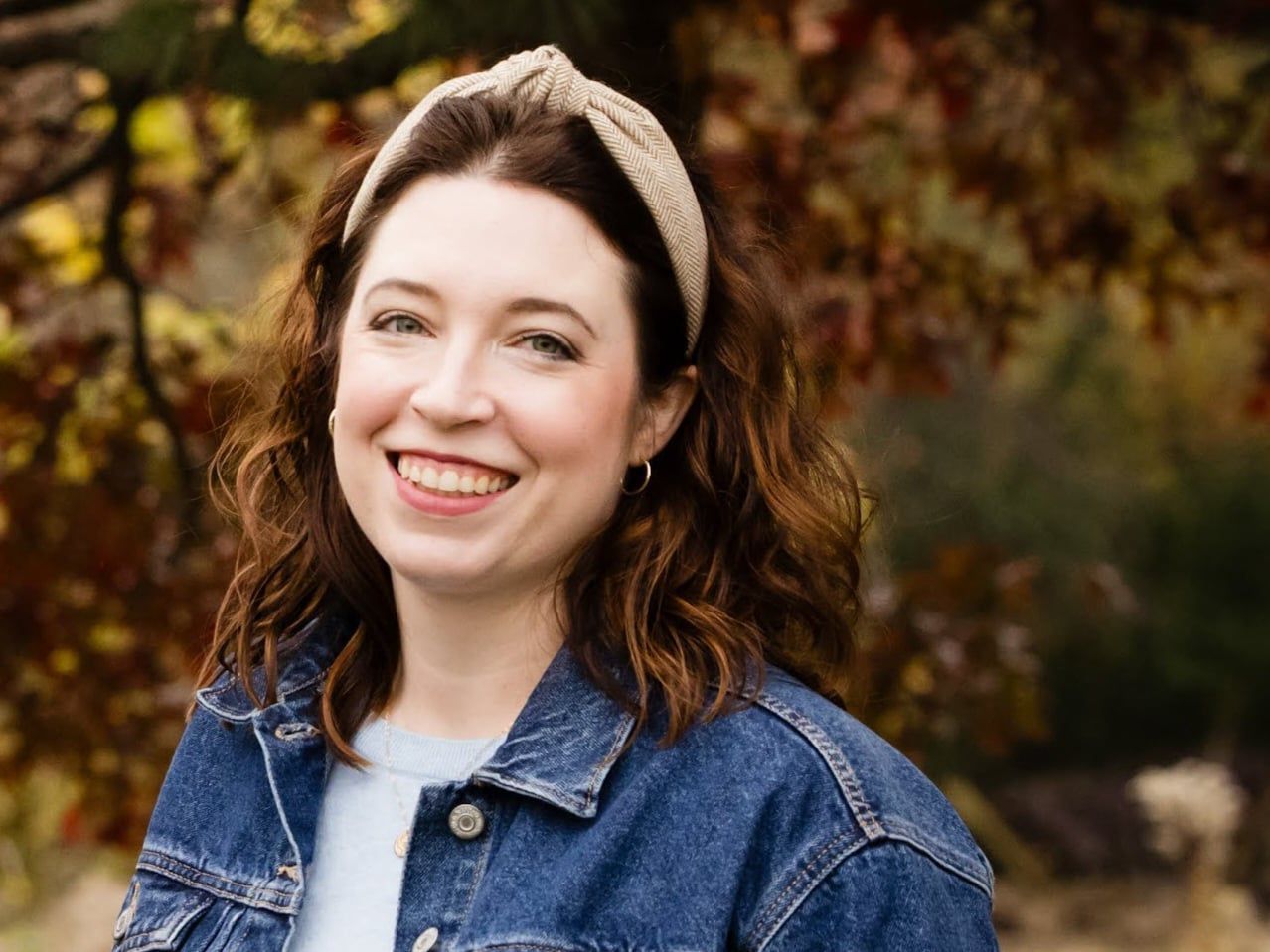 Sarah Smith in a denim jacket and knit headband outdoors with blurred autumn foliage behind them