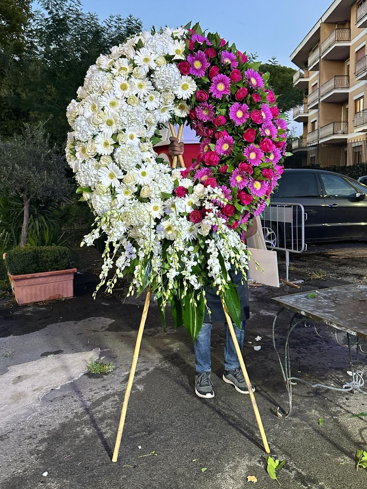 Ghirlanda floreale a forma di cuore, composta da fiori bianchi e rosa, posizionata su un supporto all'aperto, accanto a un edificio e a un'auto parcheggiata.