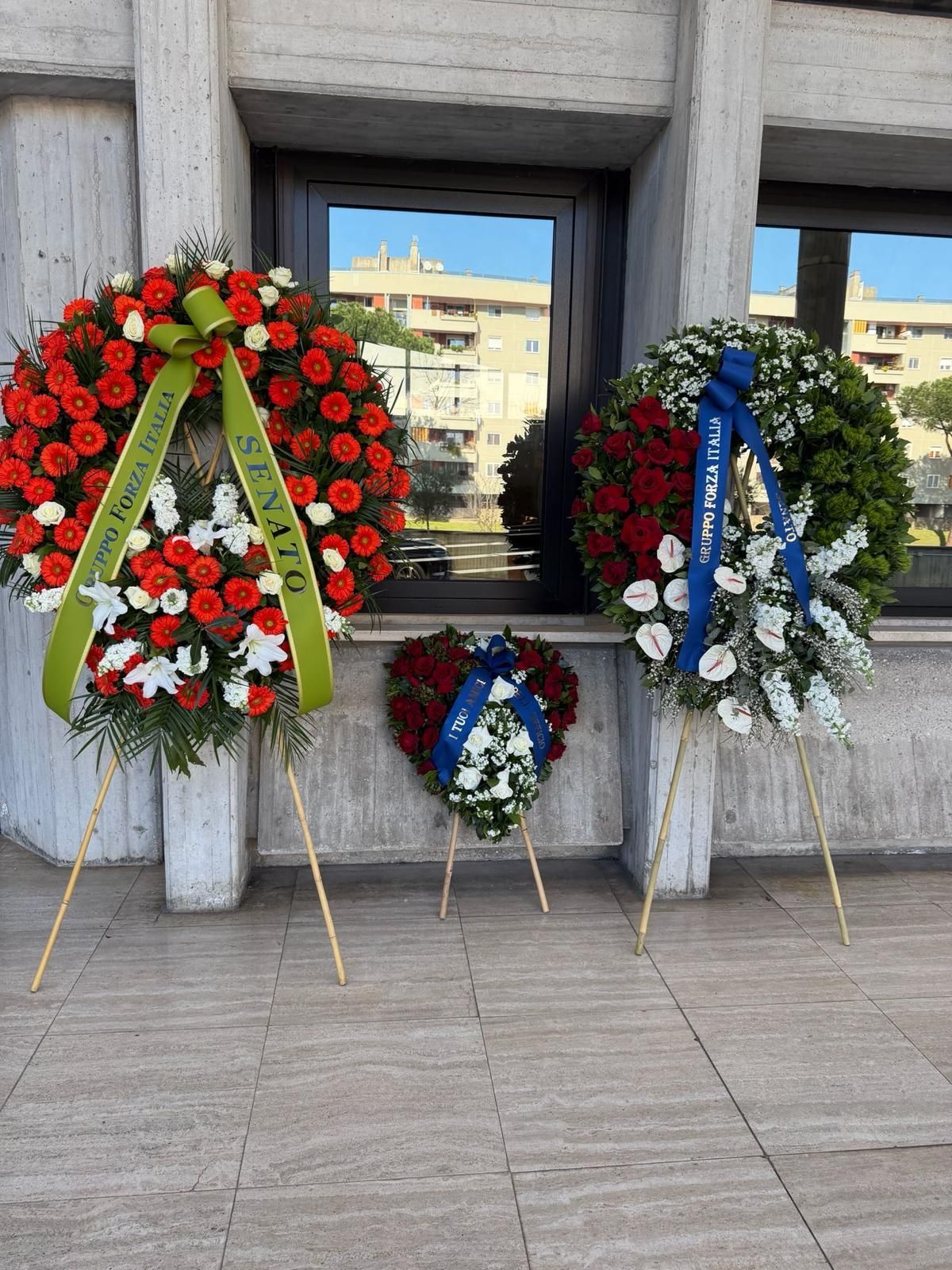 Tre corone di fiori rossi, bianchi e blu esposte davanti a un muro commemorativo in pietra.