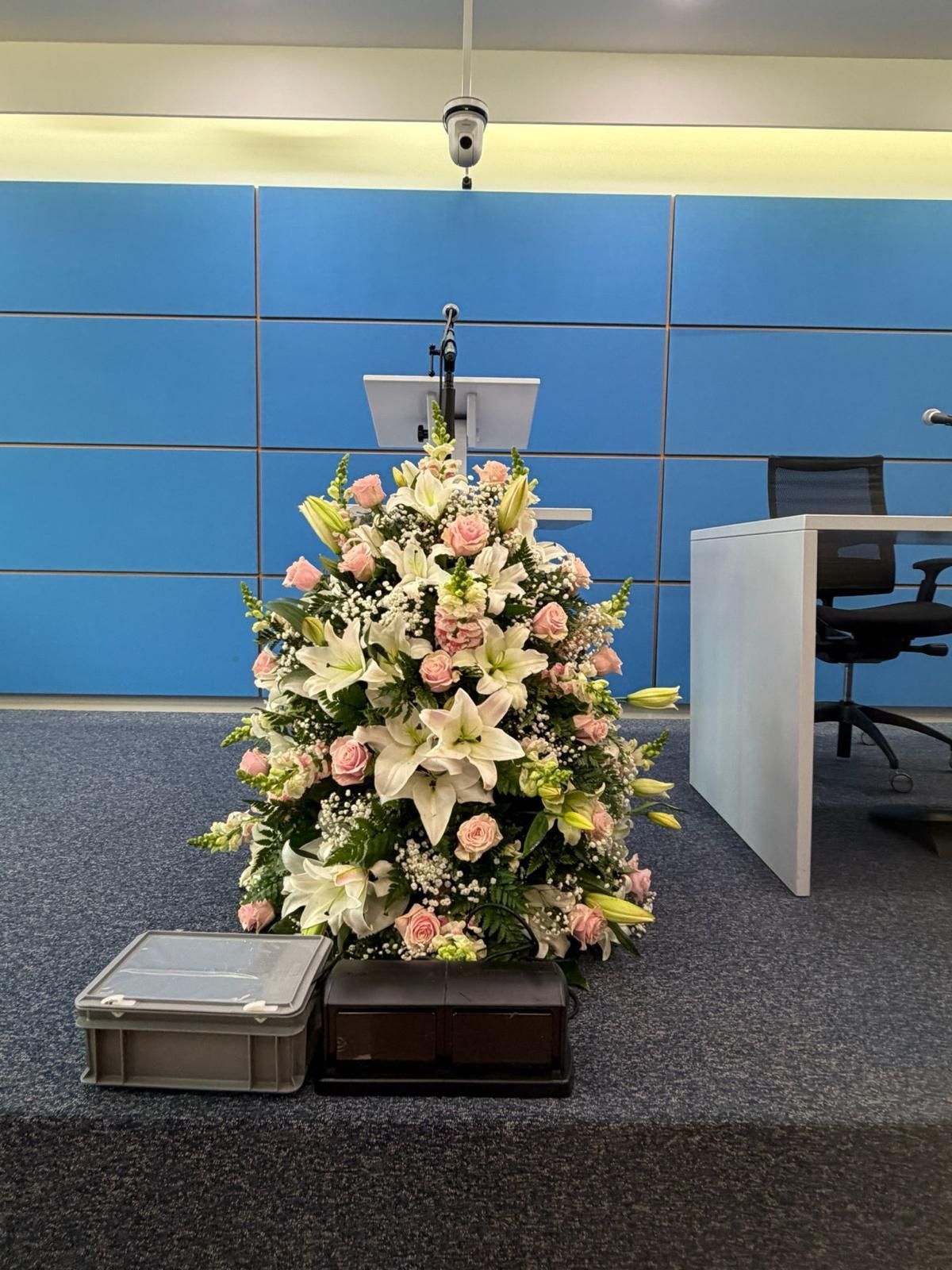 Composizione floreale funebre con fiori rosa e bianchi in una sala conferenze, accanto a due piccole valigette sul pavimento.