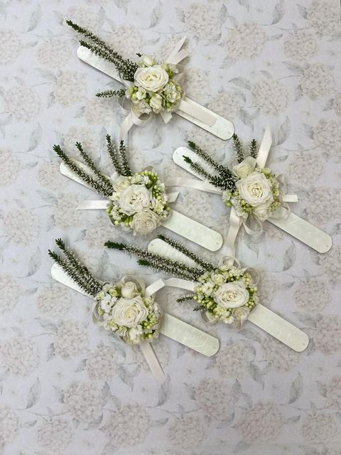 Cinque boutonnière di fiori bianchi con verde disposte su una superficie chiara con motivo decorativo.