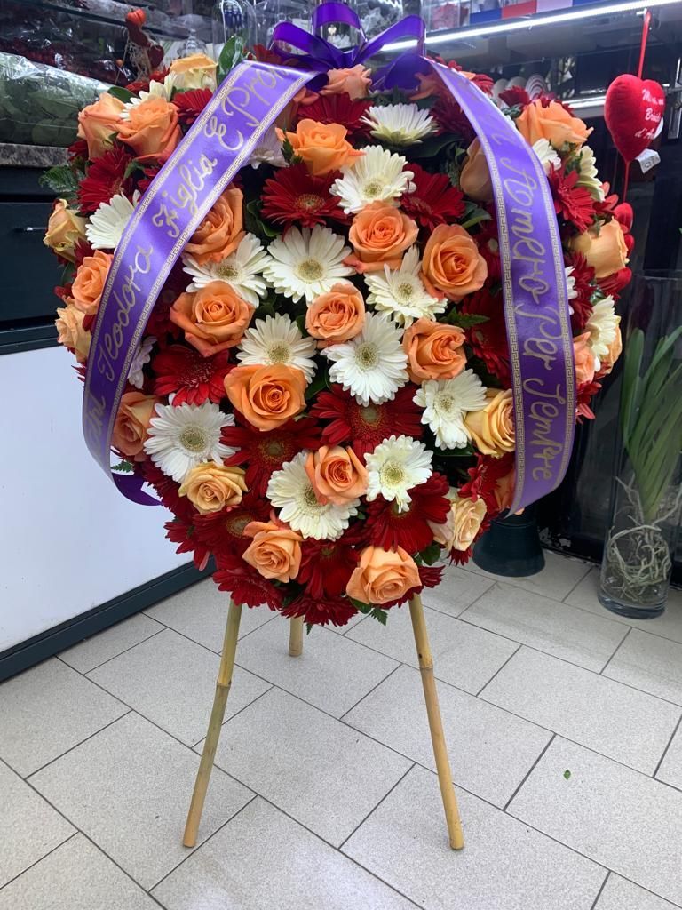 Ghirlanda floreale a forma di cuore con fiori arancioni, bianchi e rossi, legata con nastri viola su un supporto