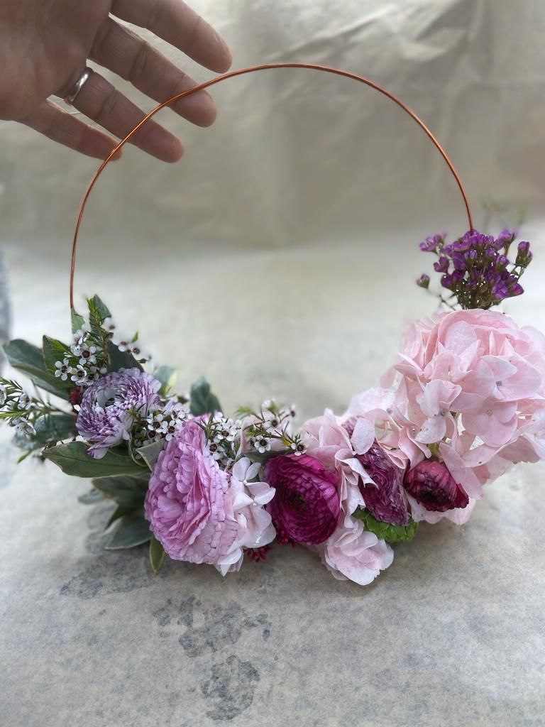 Composizione floreale ad anello realizzata a mano con fiori rosa e viola, sorretta da una mano.