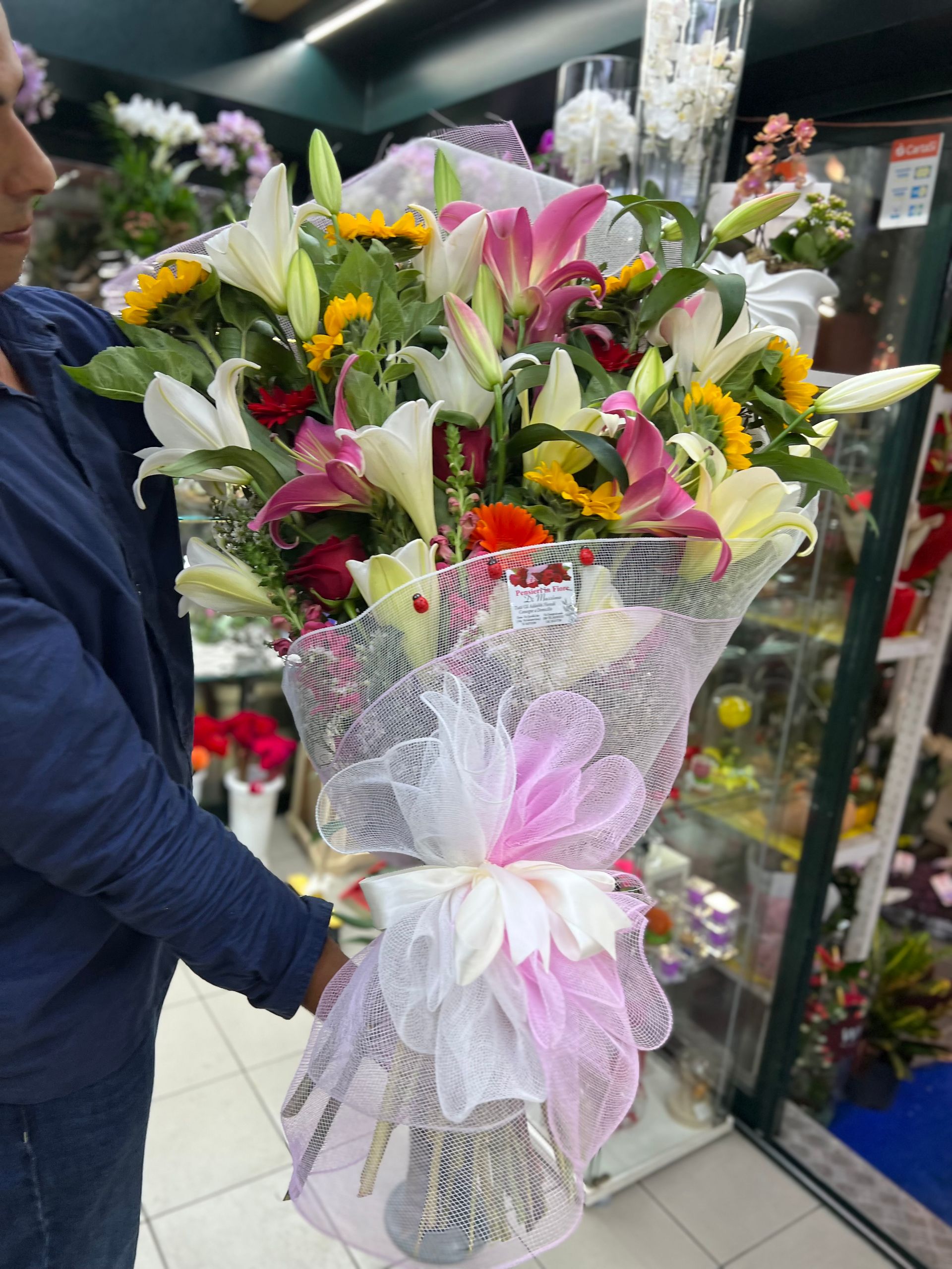 Un grande mazzo di gigli e rose avvolto in una rete bianca con un fiocco rosa, tenuto in mano in un negozio di fiori.