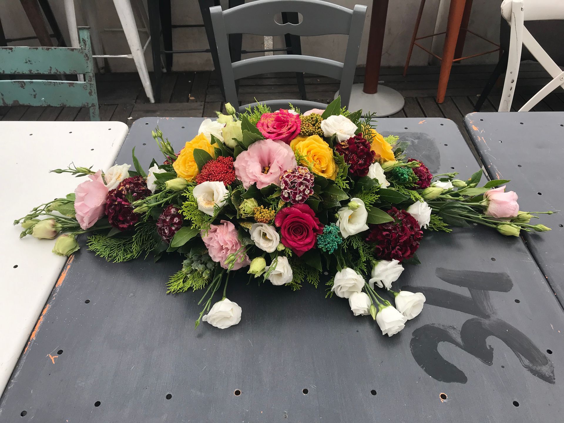 Composizione floreale colorata su un tavolo bagnato all'aperto, con fiori rosa, bianchi, gialli e rossi.