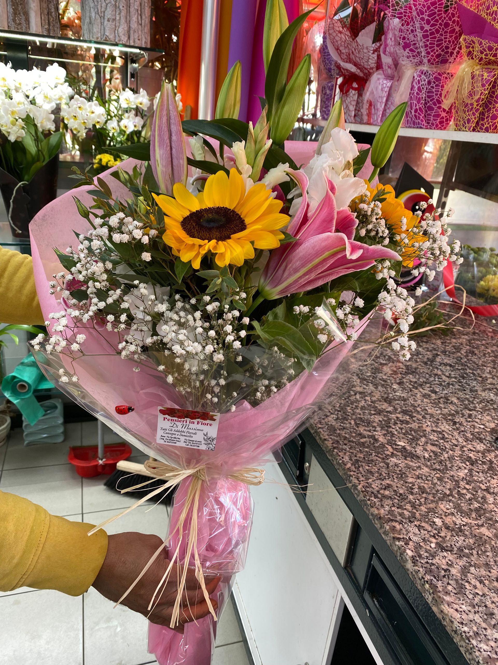 Mano che tiene un bouquet con un girasole e gigli rosa avvolti in carta rosa