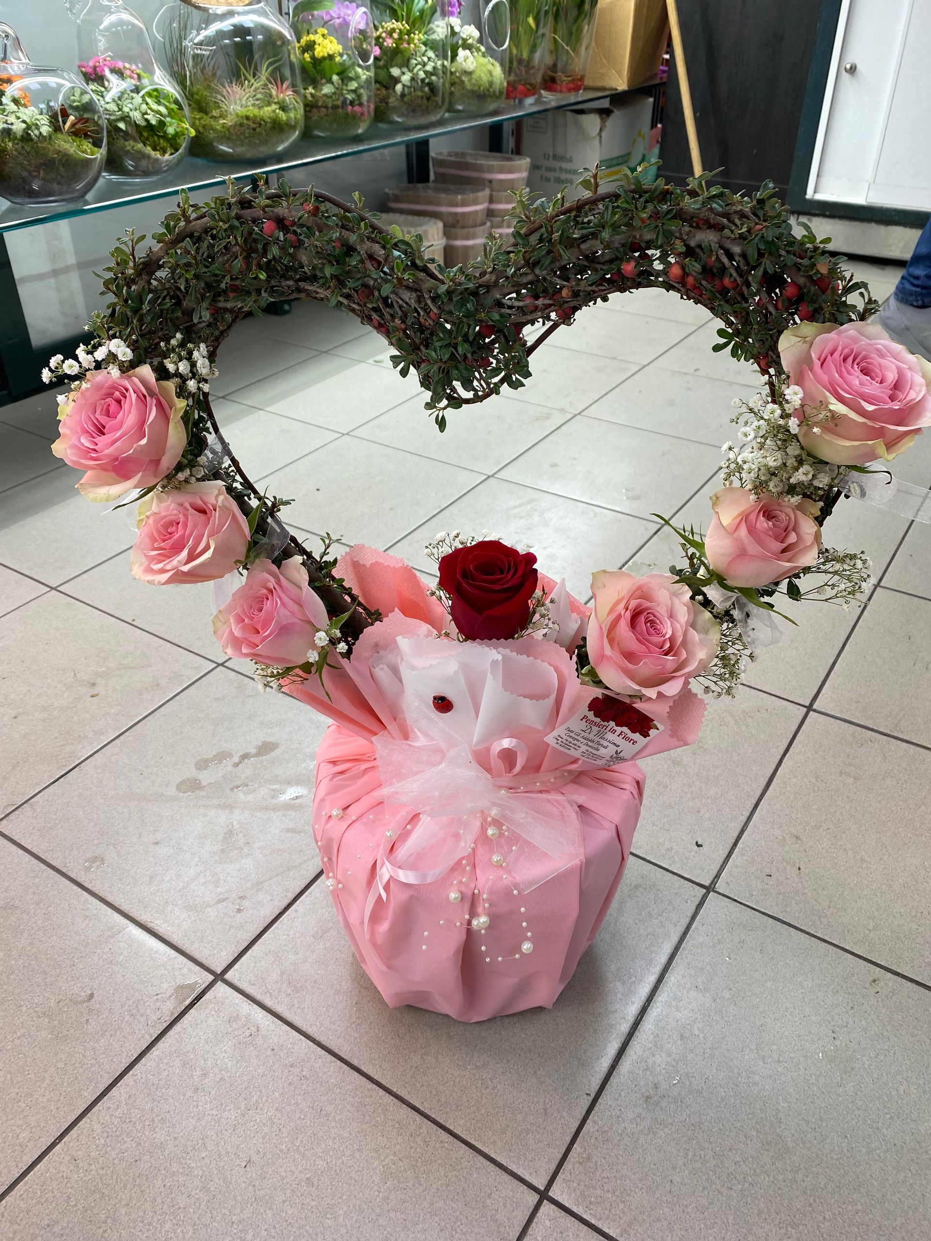 Composizione floreale a forma di cuore con rose rosa e rosse, avvolta in una carta regalo rosa, su un pavimento piastrellato.