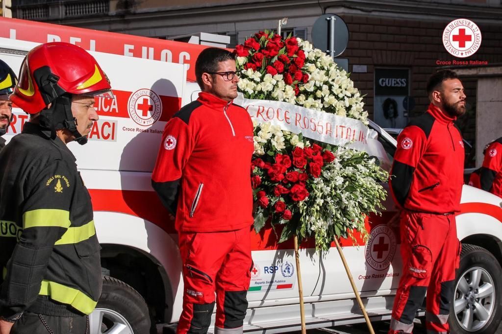 I soccorritori della Croce Rossa accanto a un camion con una grande corona di fiori, presso un memoriale allestito a bordo strada.