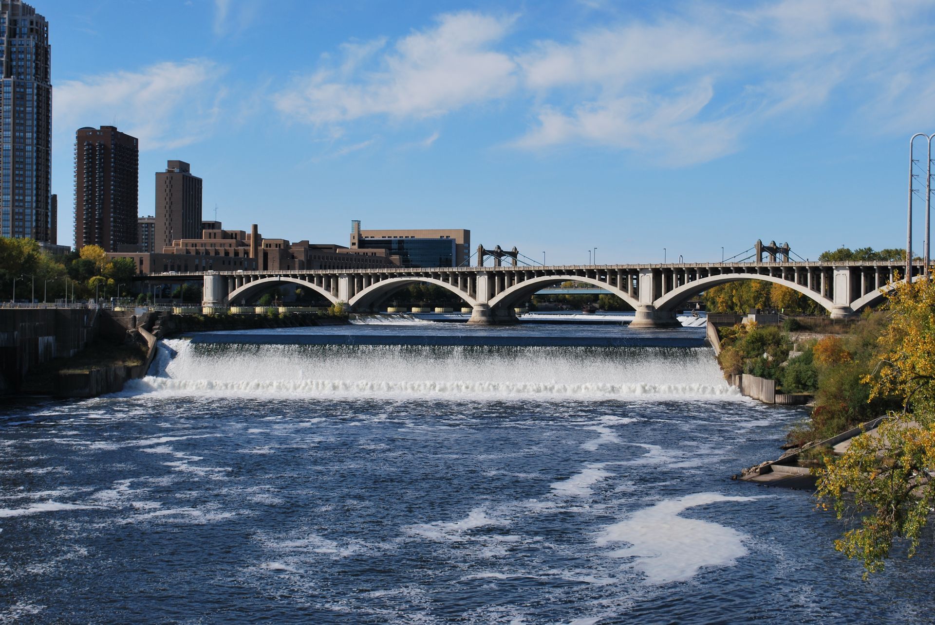 Stone Arch Bridge over a waterfall on a river, with city buildings and blue sky.