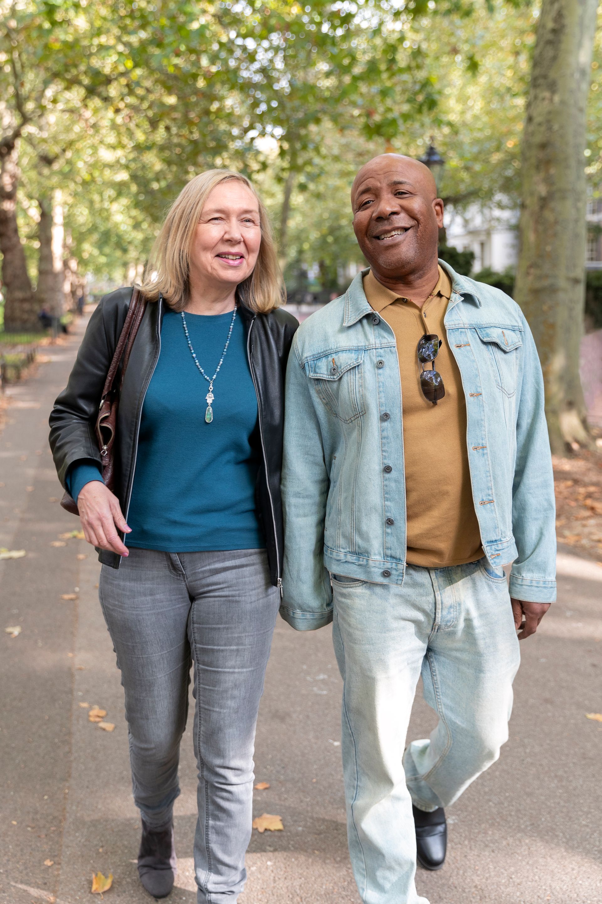 Couple walking and holding hands on a tree-lined path; they are smiling.