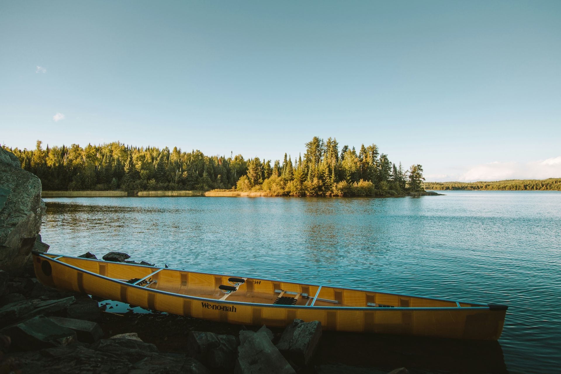 Yellow canoe rests on a rocky shore, overlooking a lake and an island with trees under a clear blue sky.