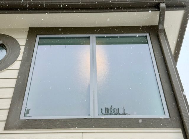 Snow falling outside a window framed by brown trim; part of a building visible.