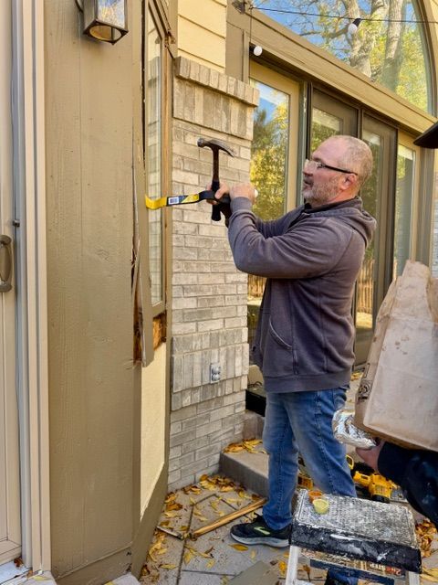 Man using a hammer, working on the side of a building next to a window; exterior shot.