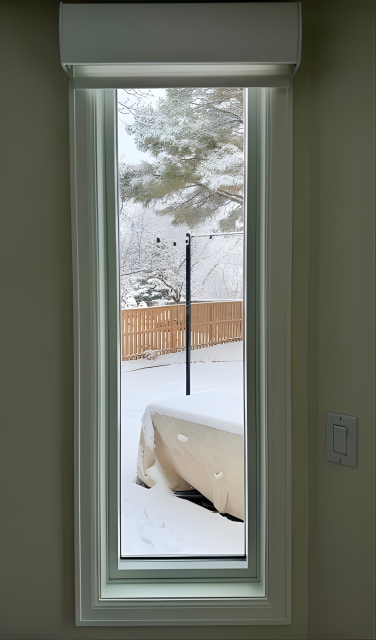 Tall, rectangular window frames a snowy backyard scene. White trim and top blind box, with an off-white wall.