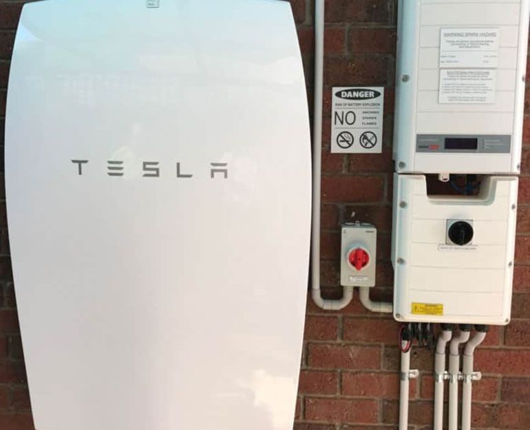 Tesla Powerwall battery mounted on brick wall next to control panel with wiring, 'Danger' sign for electrical hazards.