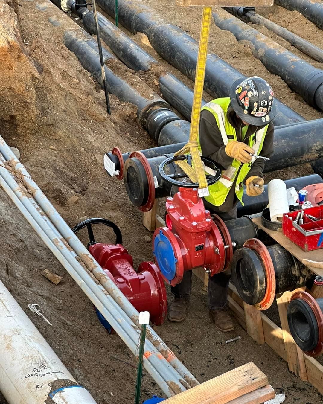 A construction worker in safety gear measures a large red valve between black pipes at an excavation site.