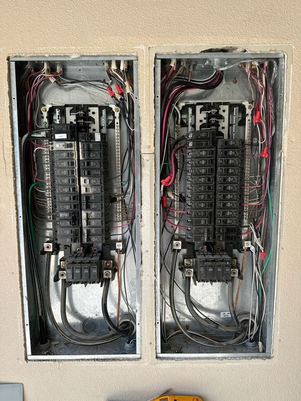 Two open electrical panels with numerous wires of different colors connected to circuit breakers inside a wall.