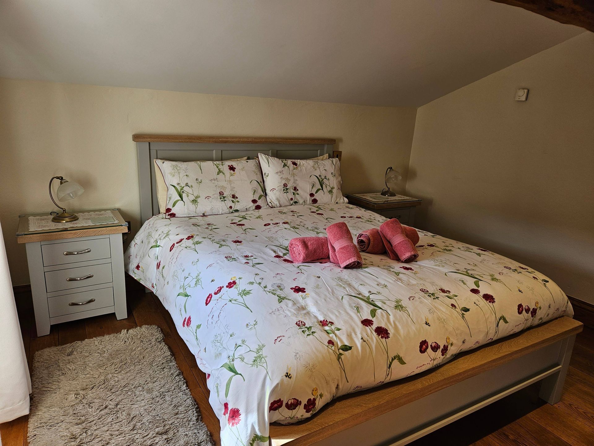 Bedroom with a bed, two nightstands, a rug, and floral bedding. Pink towels are on the bed.