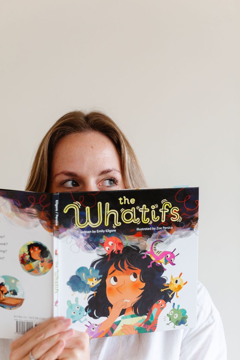 Person peeks over a colorful “The Whatifs” book cover, holding it up indoors against a plain wall.