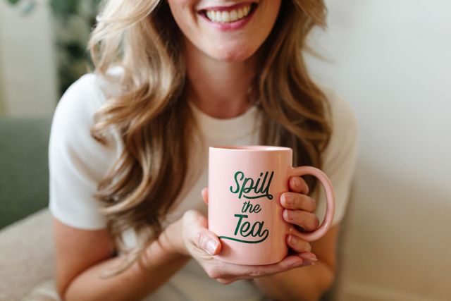 Woman holding a pink mug that says “Spill the Tea” and smiling indoors