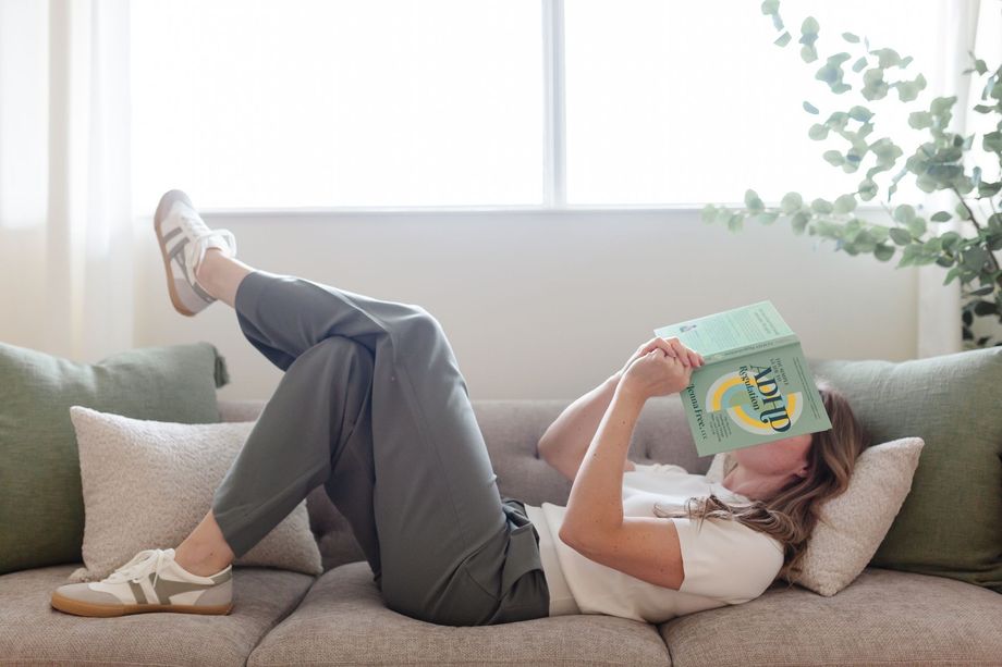 Person relaxing on a beige couch, reading a book with feet up, in a bright living room