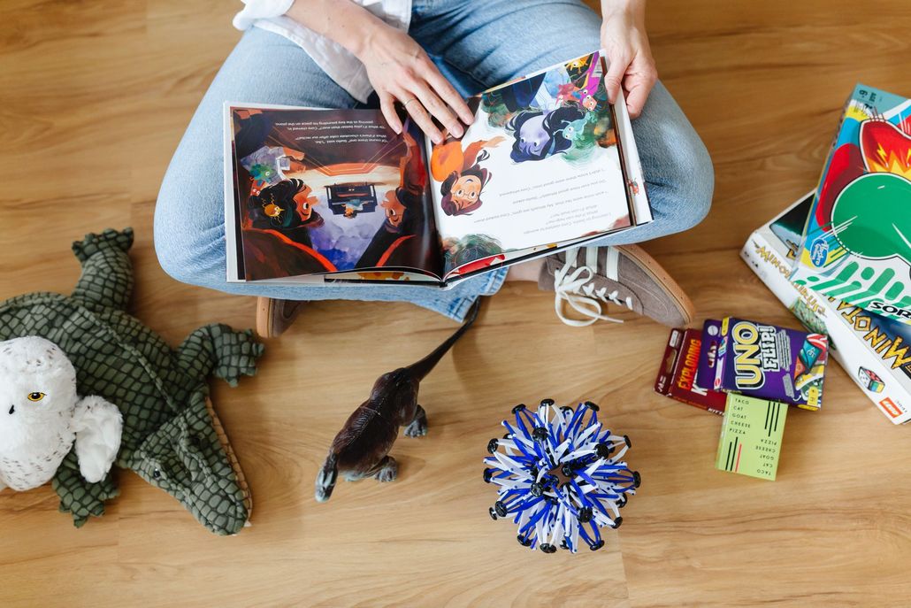 Person sitting on a wooden floor reading a colorful book with toys and wrapped gifts nearby