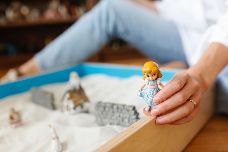 Hand placing a small doll in a sandbox with miniature stone walls and figures