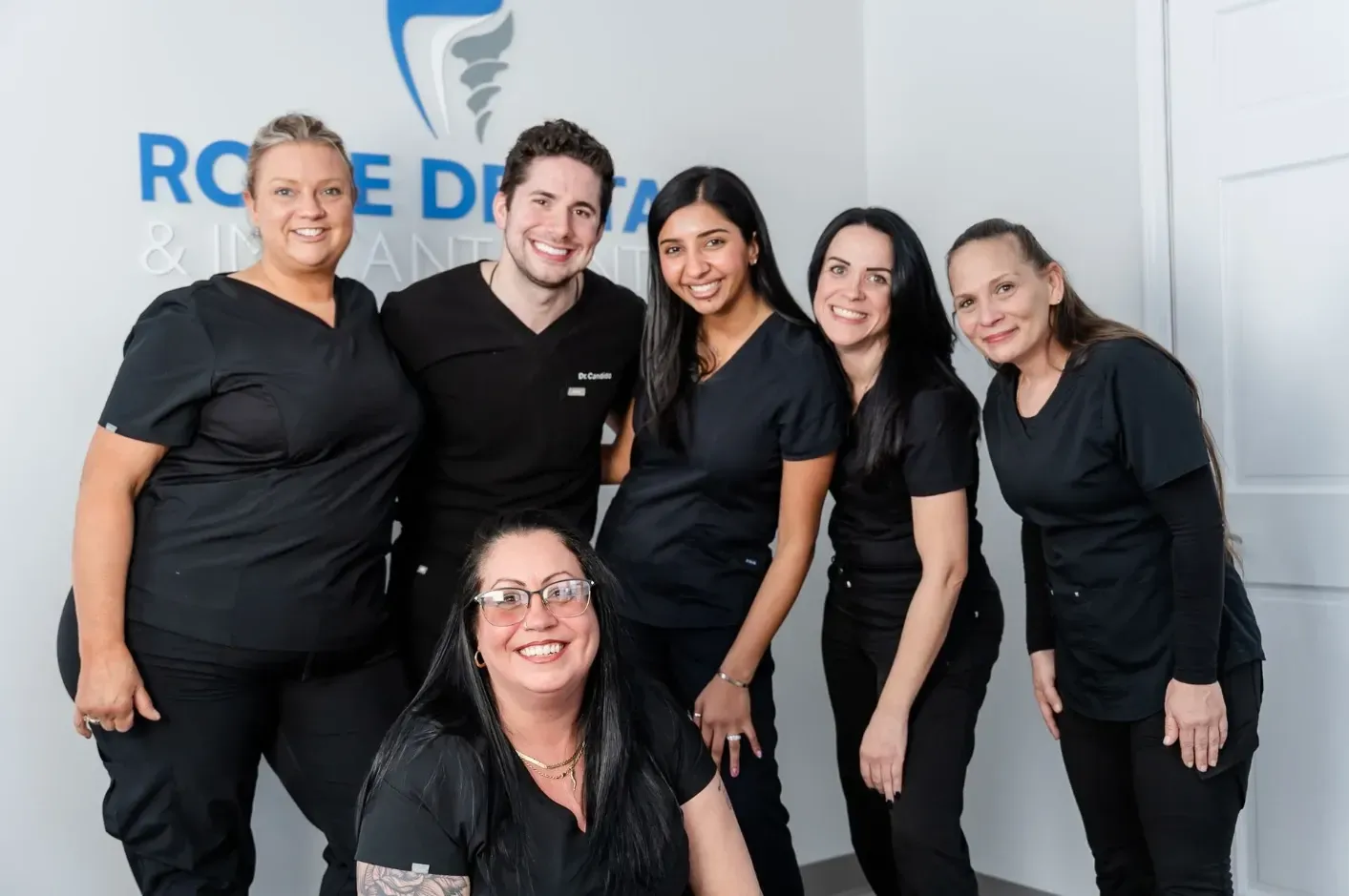 Group of six dental professionals smiling in front of a clinic logo. All are wearing black scrubs.