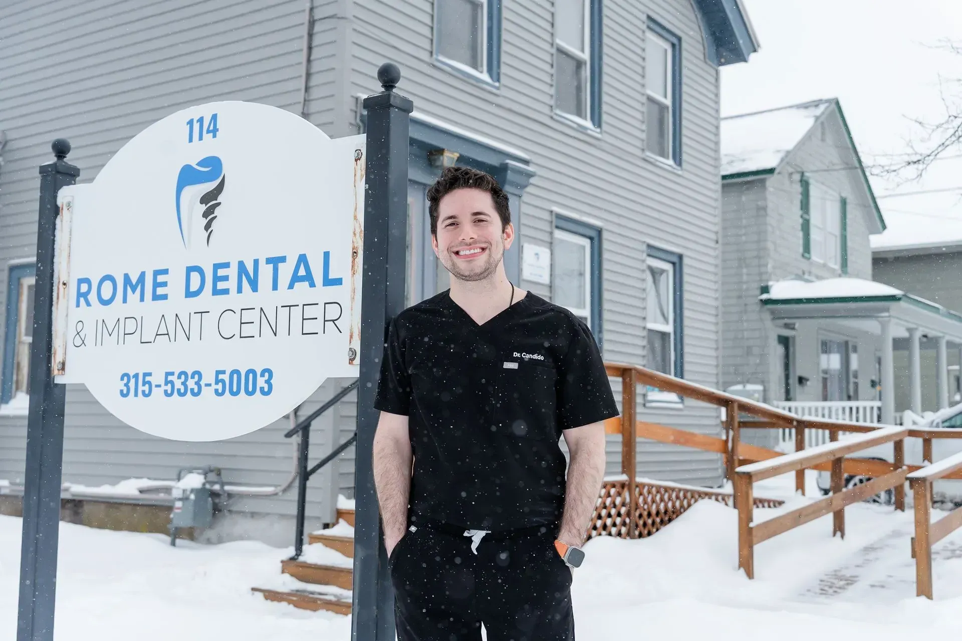 Man in black scrubs smiles in front of Rome Dental & Implant Center in snowy weather.