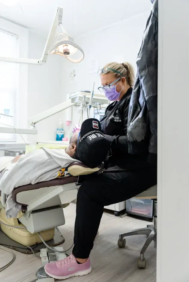 Dentist in black scrubs, mask, and pink shoes working on a patient in a dental chair.